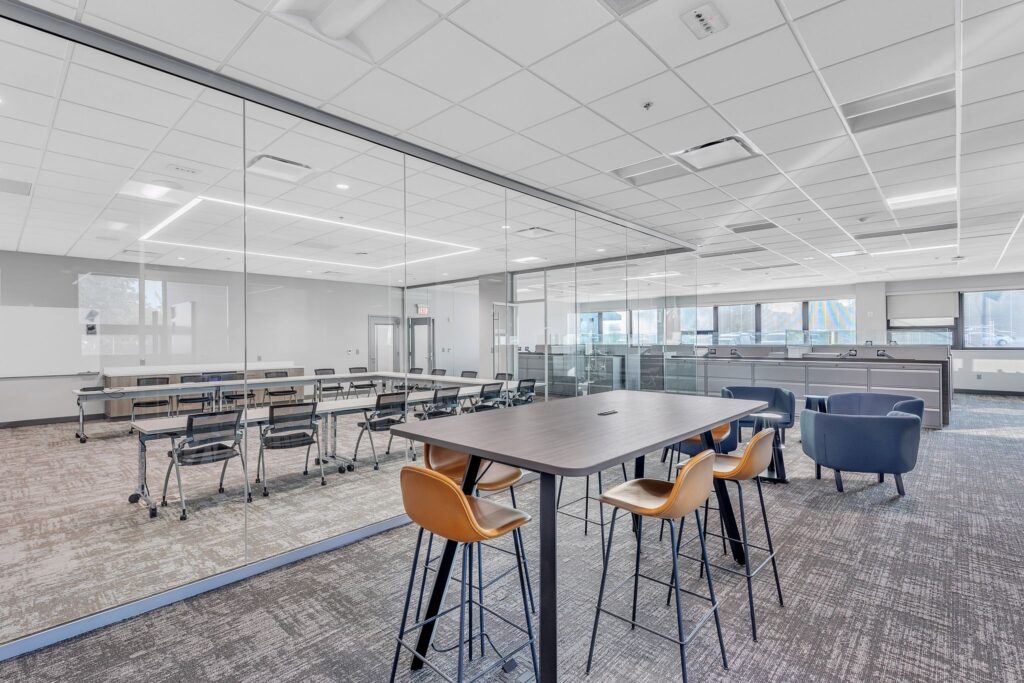 A modern office space featuring a glass-enclosed conference room with tables and chairs, and a common area with high-top seating and blue lounge chairs.