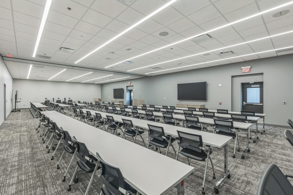 A large conference room with rows of tables and chairs facing two wall-mounted televisions.
