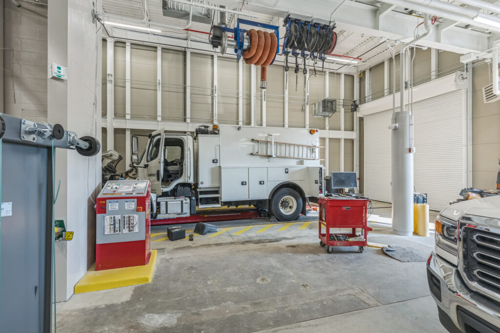 A utility truck parked inside a maintenance bay, with a red diagnostic control station and a mobile tool cart nearby.