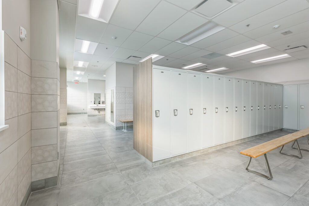 A locker room featuring a long row of white lockers, a wooden bench, and an open doorway leading to a washroom area.