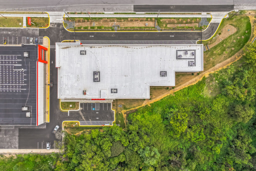 Aerial view of a commercial building with a large flat roof, adjacent parking lot, and surrounding wooded area.