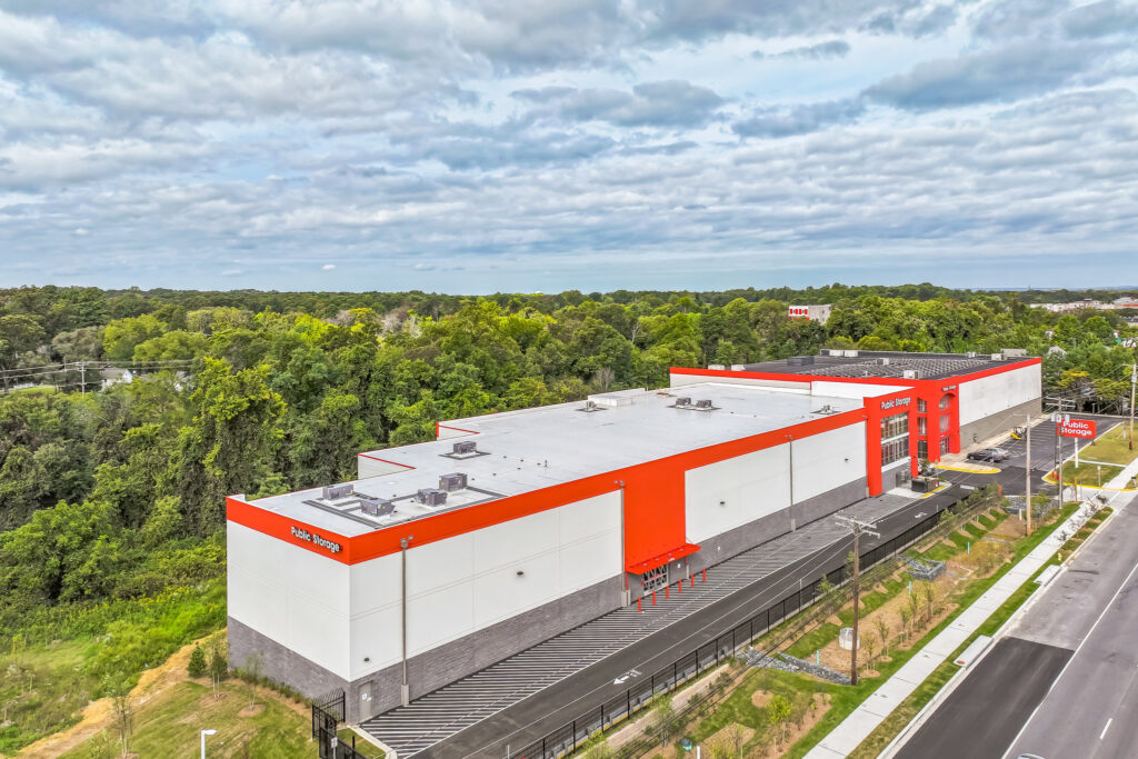 An aerial view of a Public Storage building with a grey and white exterior and orange trim, situated next to a treed area and a road.