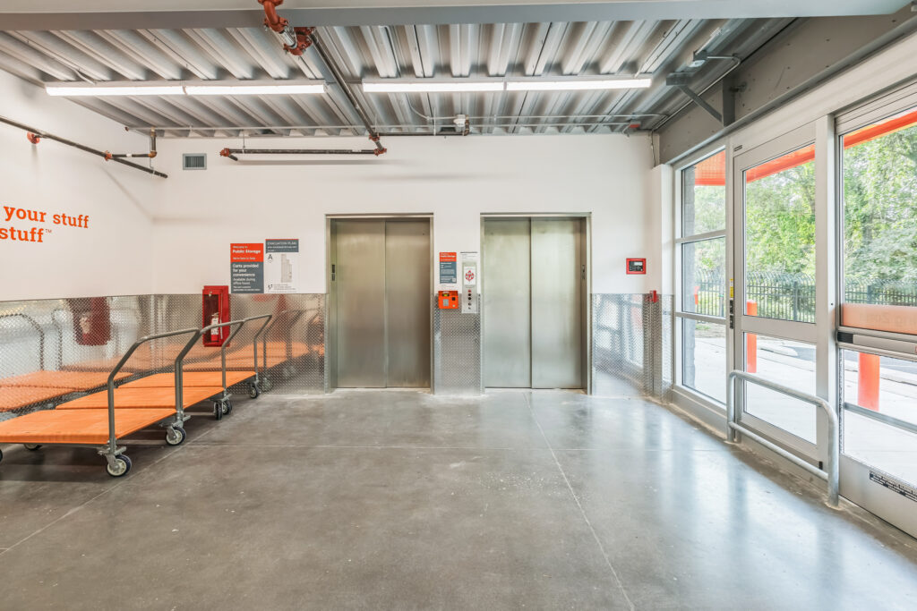 Two stainless steel elevators in a storage facility, with orange rolling flatbed carts parked to the left and a glass entrance door to the right.