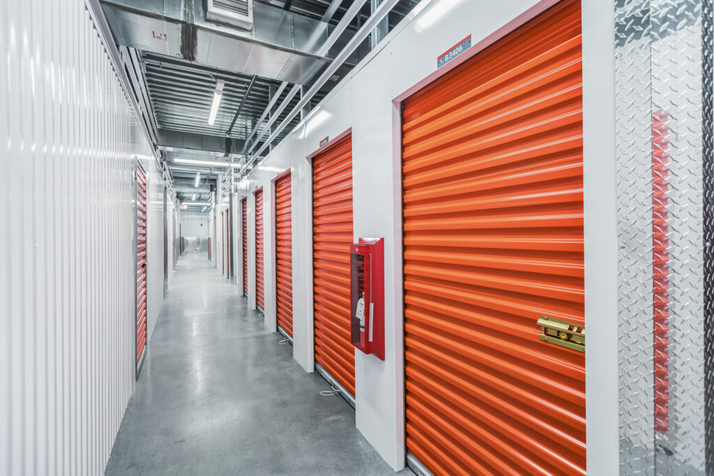 A hallway in a self-storage facility featuring a row of individual storage units with bright orange roll-up doors.