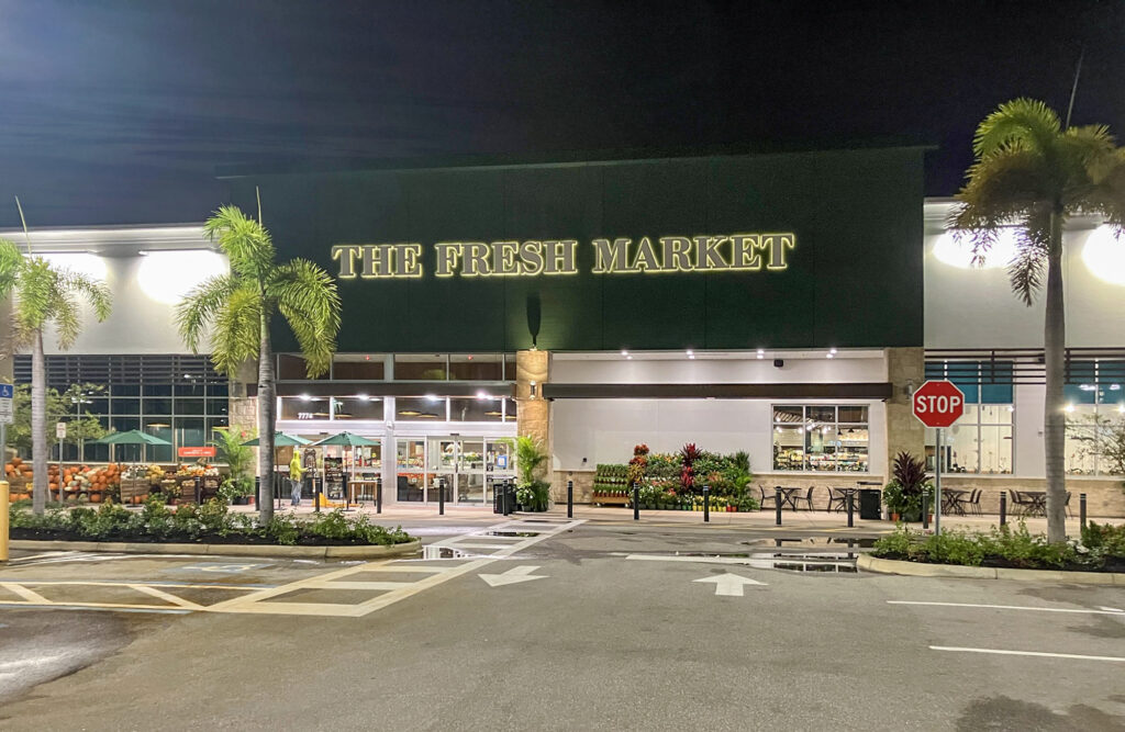 The exterior of The Fresh Market grocery store at night, with palm trees and a parking lot in the foreground.