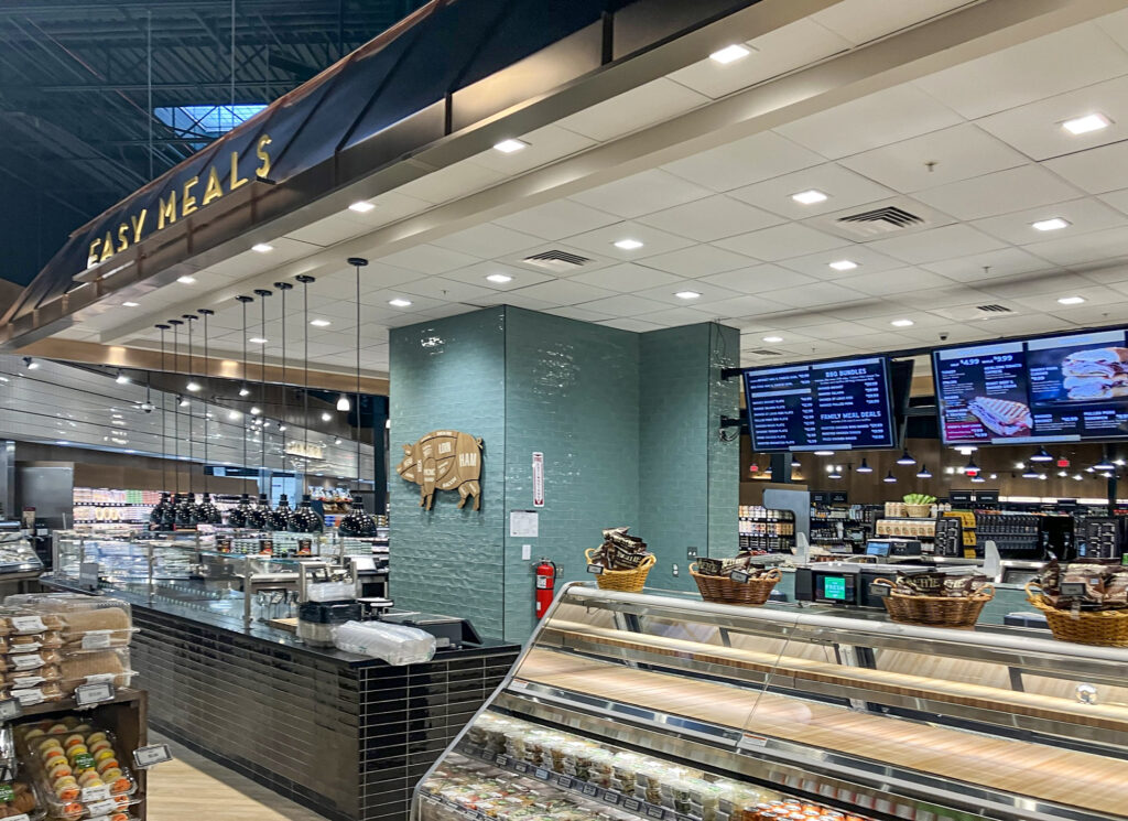 Grocery store deli counter with an “Easy Meals” sign, a pig meat-cut diagram on a green tile wall, and digital menu boards displaying meal deals.