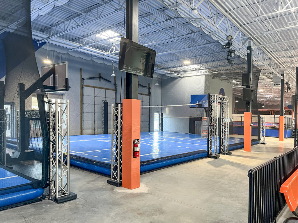 A multi-purpose indoor sports court with blue padded flooring, basketball hoops, and a volleyball net surrounded by metal truss structures.