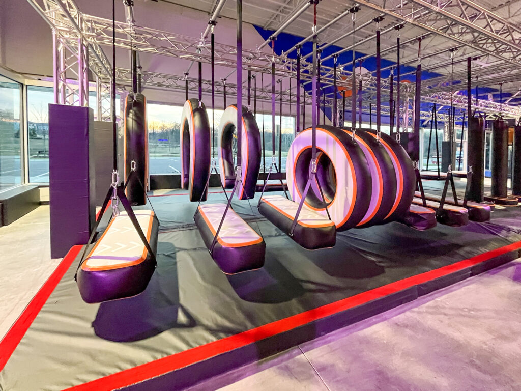 An indoor obstacle course featuring a series of hanging padded rings and balance beams suspended from an overhead metal truss.