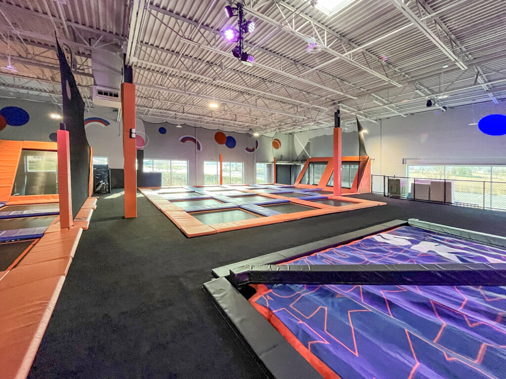 An indoor trampoline park with multiple jumping surfaces, black flooring, and orange safety padding.
