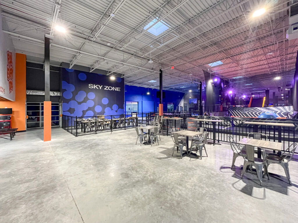 An indoor Sky Zone trampoline park featuring multiple seating areas with metal tables and chairs on a concrete floor.