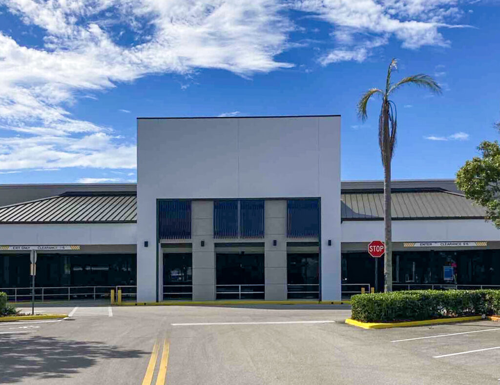 Exterior of a commercial building with large glass windows and a central entrance, with a palm tree and stop sign in the foreground.