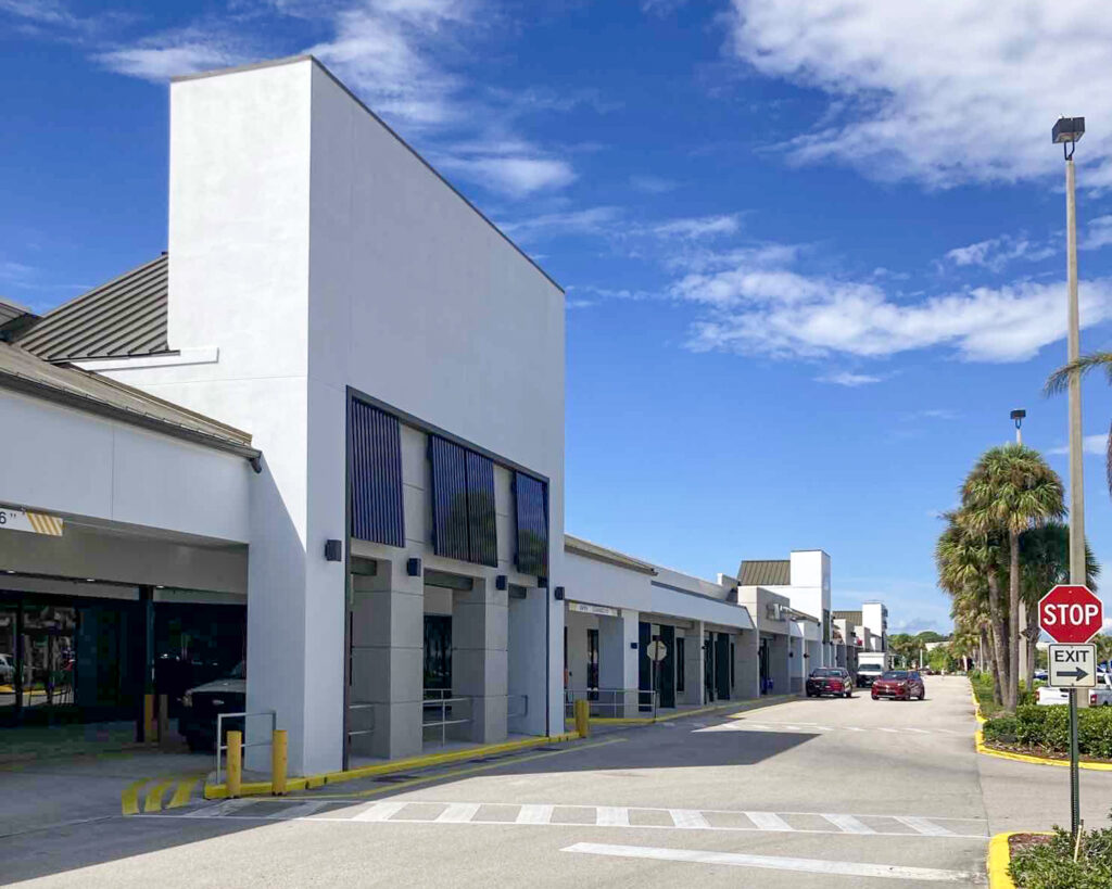 A strip mall parking area with white storefronts, a covered walkway, and a stop sign at an exit.