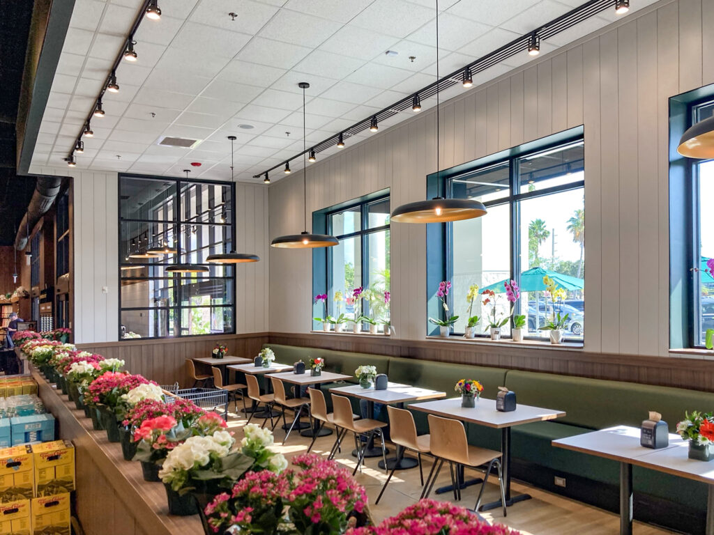 Rows of potted pink and white flowers line the dining area of a restaurant, featuring wooden tables with chairs along a green bench seat and large windows.