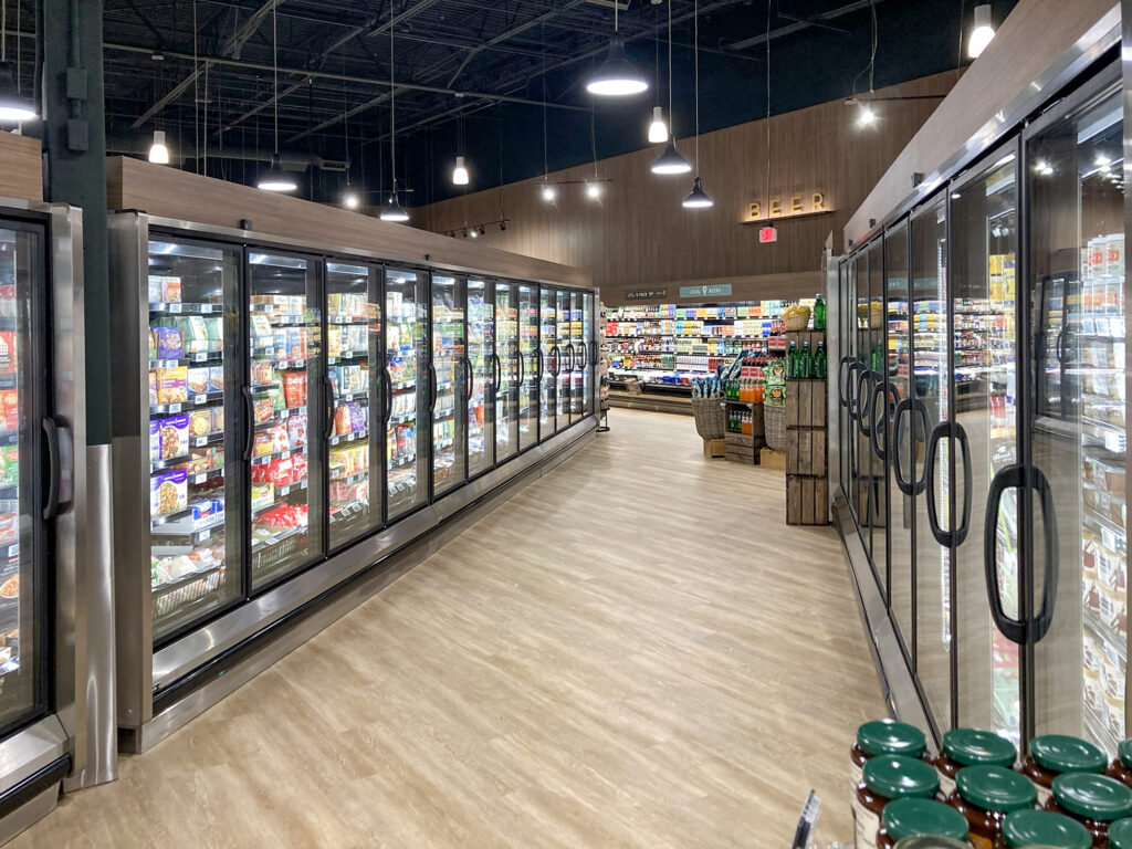 A long aisle in a grocery store featuring two rows of glass-front freezer cases and a beer section in the distance.