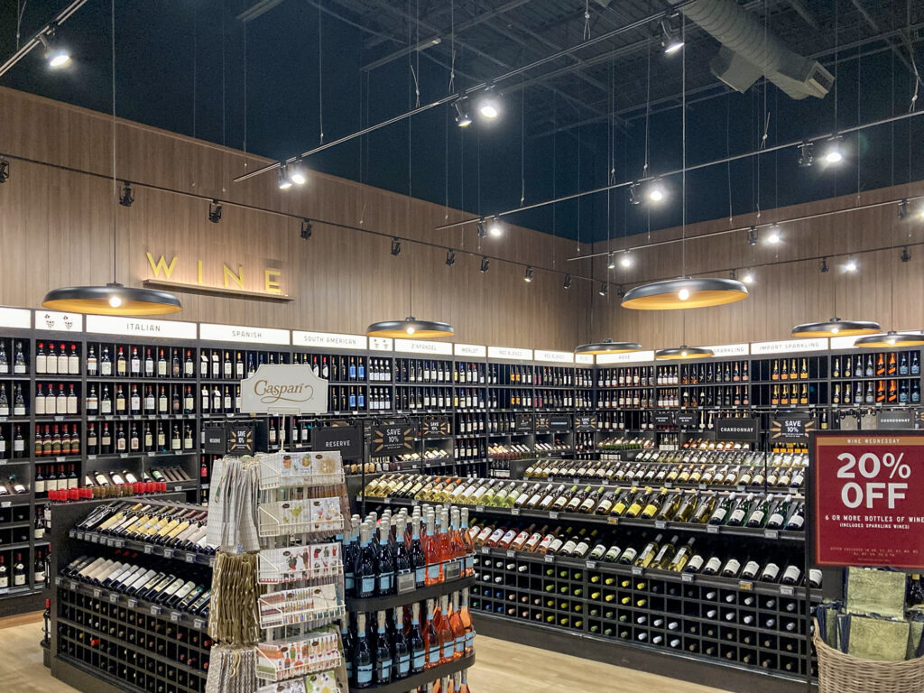 A wide aisle in a wine shop with shelves organized by region and variety, featuring a sign advertising a 20% discount on six or more bottles.