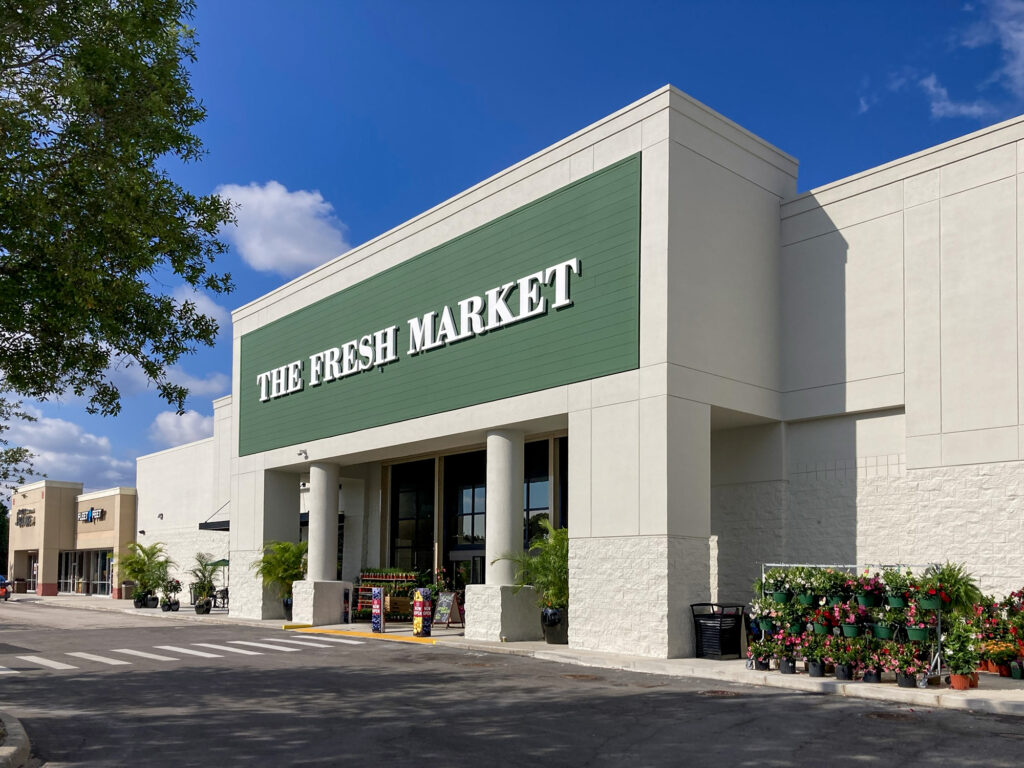 The Fresh Market grocery store storefront under a blue sky, with potted flowers displayed outside the entrance.