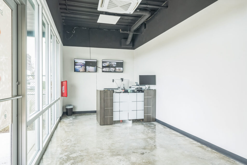A reception desk with a computer and two wall-mounted security monitors in a room with white walls, a black ceiling, and a concrete floor.