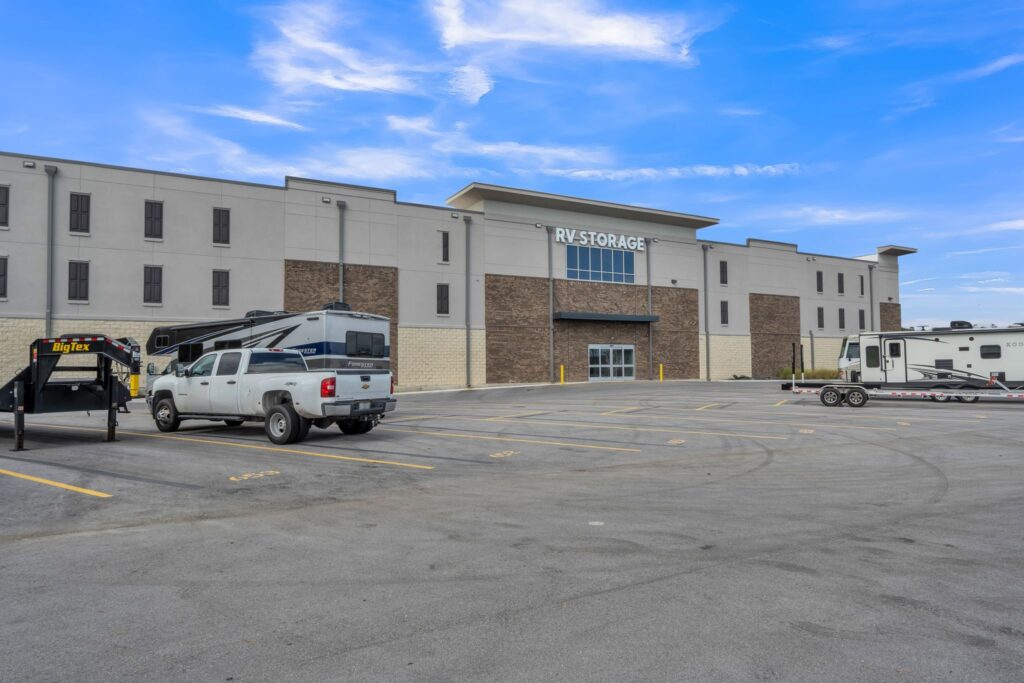 An RV storage facility building with a large parking lot containing two parked RVs and a flatbed trailer.