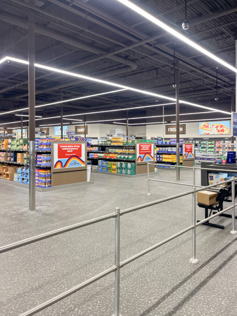 Interior of an Aldi grocery store with aisles of products, metal railings in the foreground, and signs for dairy and frozen sections.
