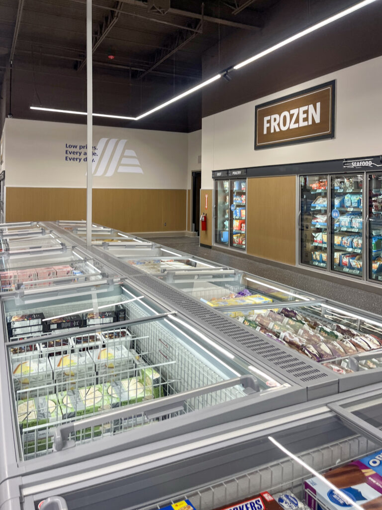 The frozen food aisle of a grocery store with chest freezers in the foreground and glass-door refrigerated cases against the back wall.