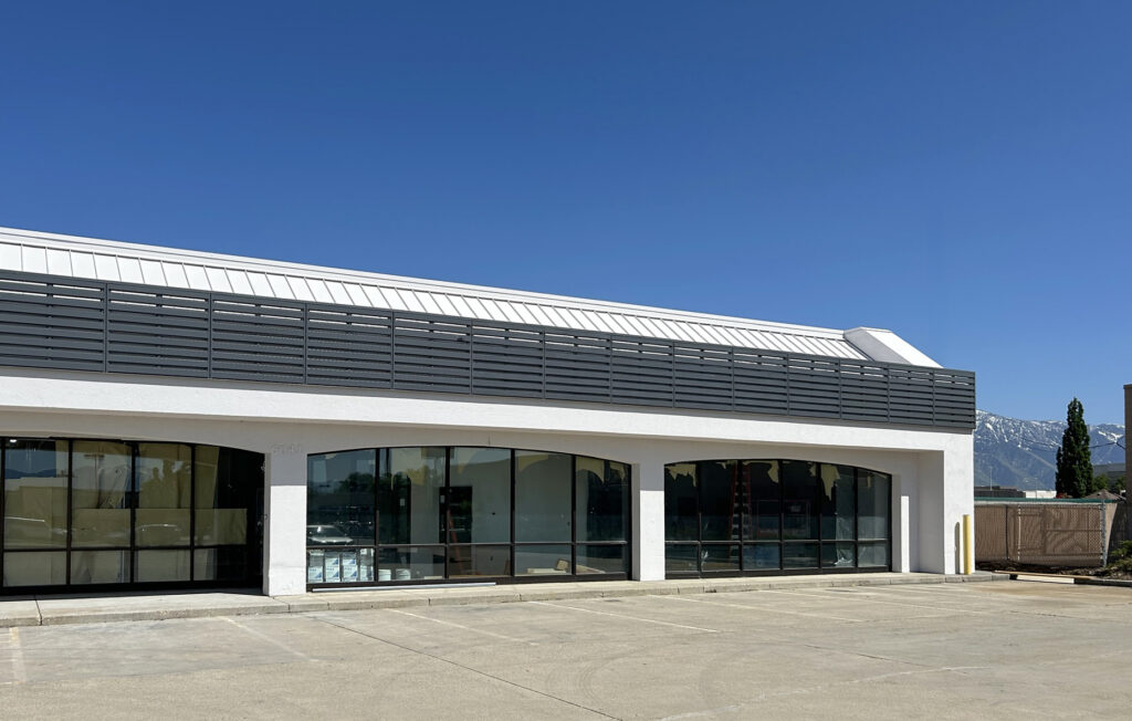 A one-story commercial building with large glass storefronts and a dark gray metal facade against a clear blue sky.