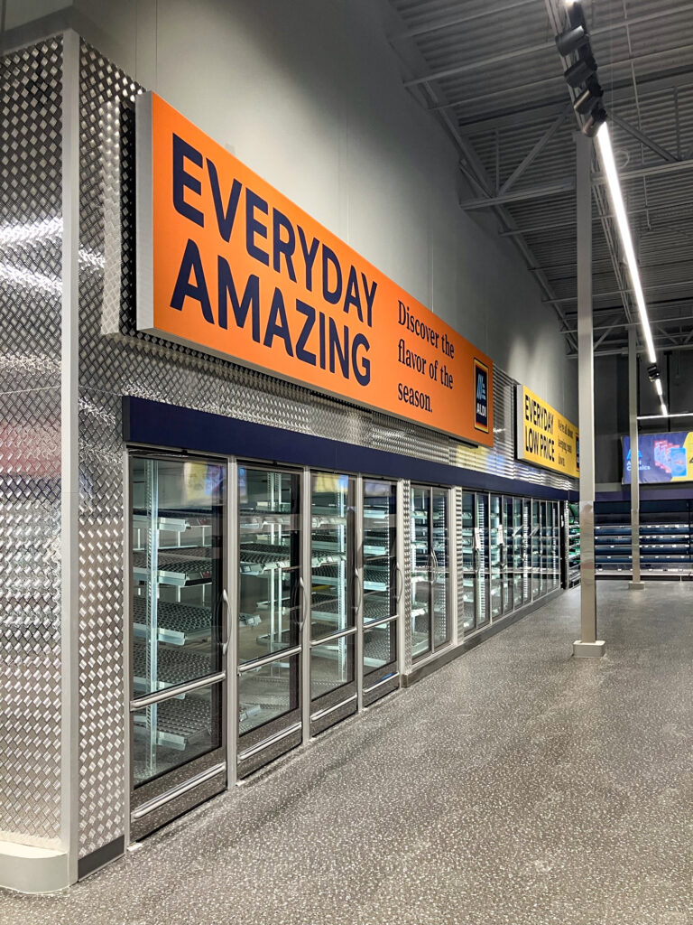 A row of empty refrigerated display cases in an Aldi grocery store under a sign that reads “Everyday Amazing.”