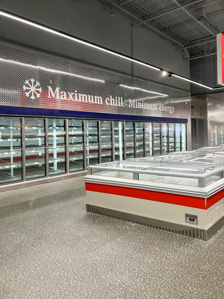 Empty glass-door freezer cases with the text “Maximum chill. Minimum energy” displayed on the diamond-plate wall above.