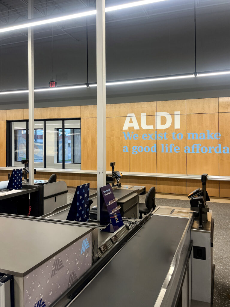 Checkout counters at an Aldi store, with the brand’s name and mission statement displayed on a wood-paneled wall.