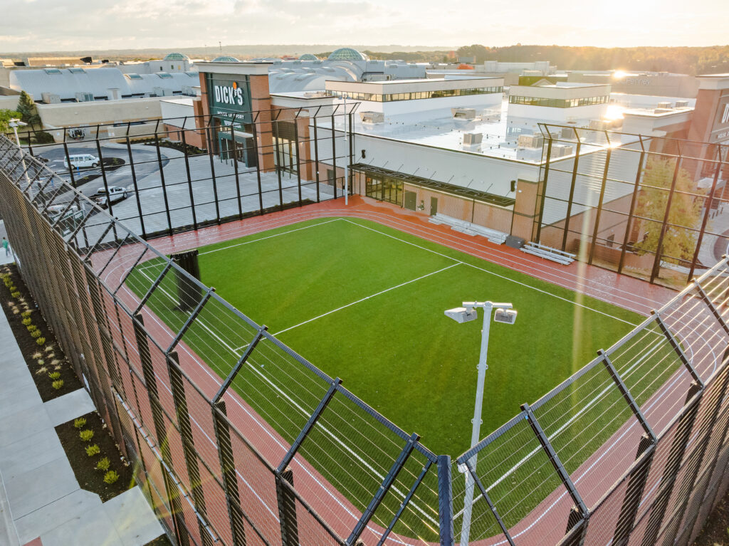 Aerial view of an enclosed artificial turf athletic field next to a large shopping mall with a Dick’s Sporting Goods store.
