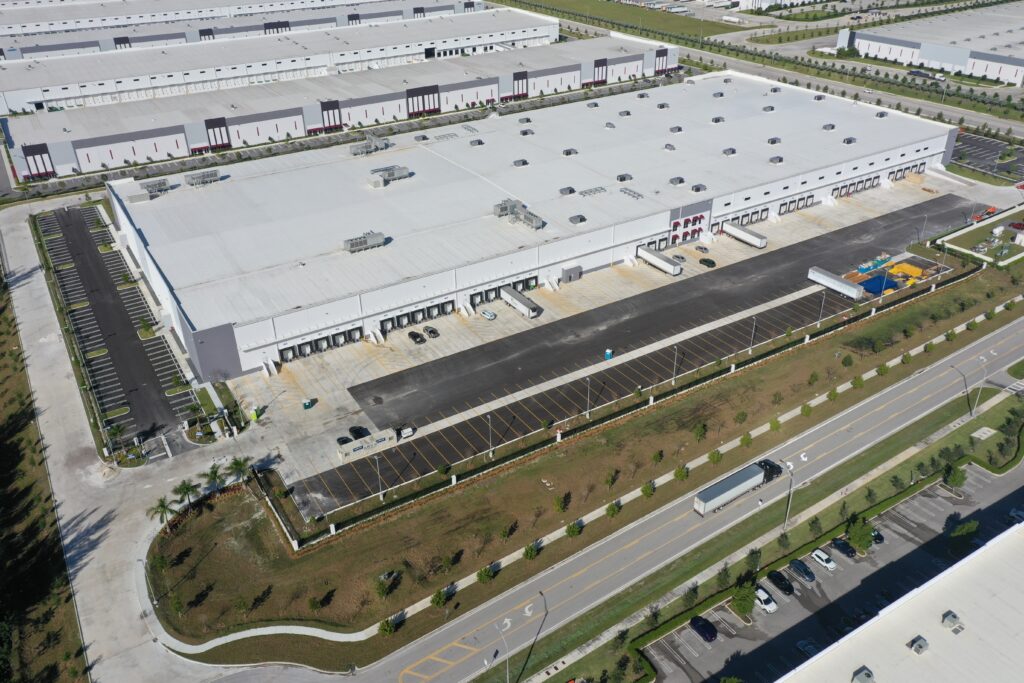Aerial view of a large industrial warehouse complex with multiple loading docks, adjacent parking lots, and a road with a moving semi-truck.