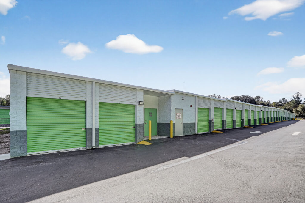 A long, one-story self-storage facility featuring a row of green roll-up metal doors and an asphalt driveway under a blue sky.