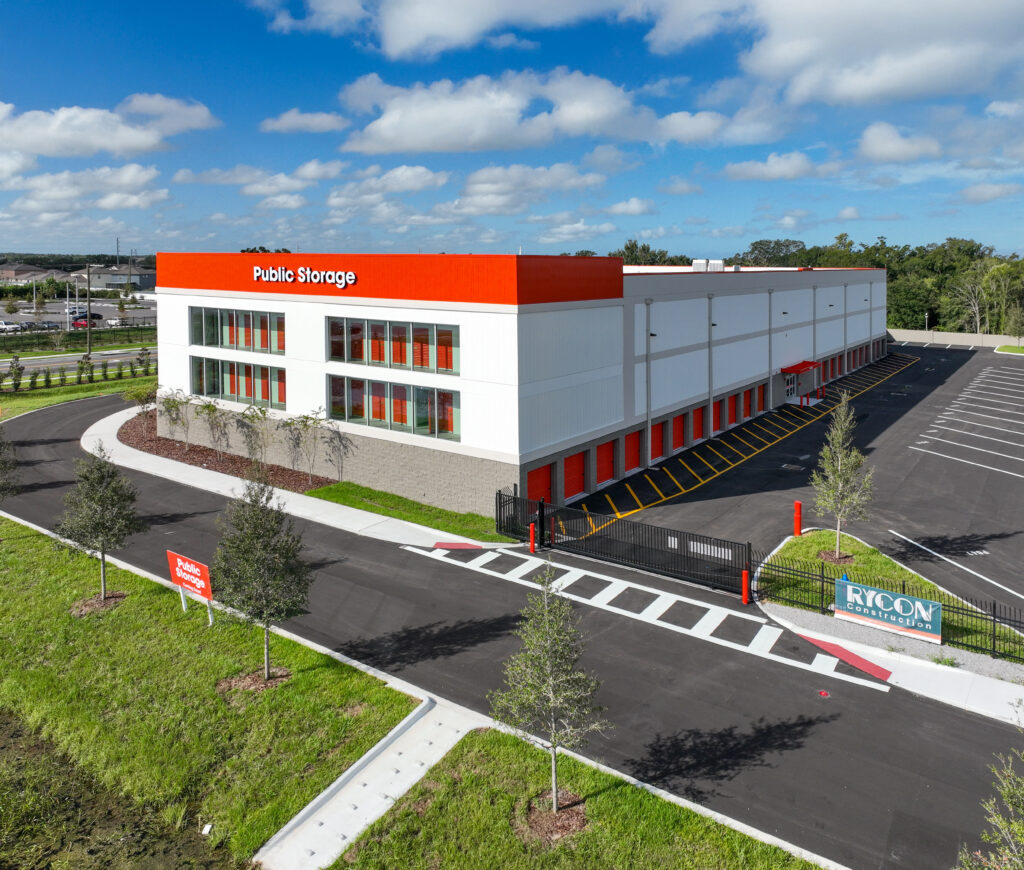 Aerial view of a Public Storage building with orange and white exterior, a gated driveway, and a Rycon Construction sign on the lawn.