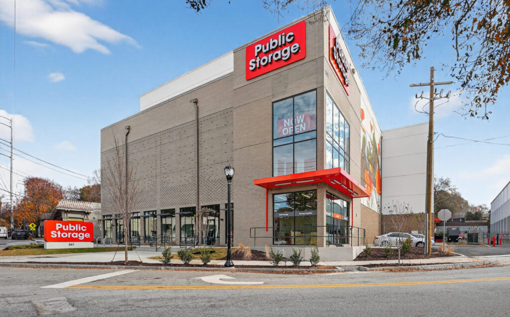 A multi-story Public Storage facility with a “Now Open” sign on the corner glass windows and a freestanding sign near the street.