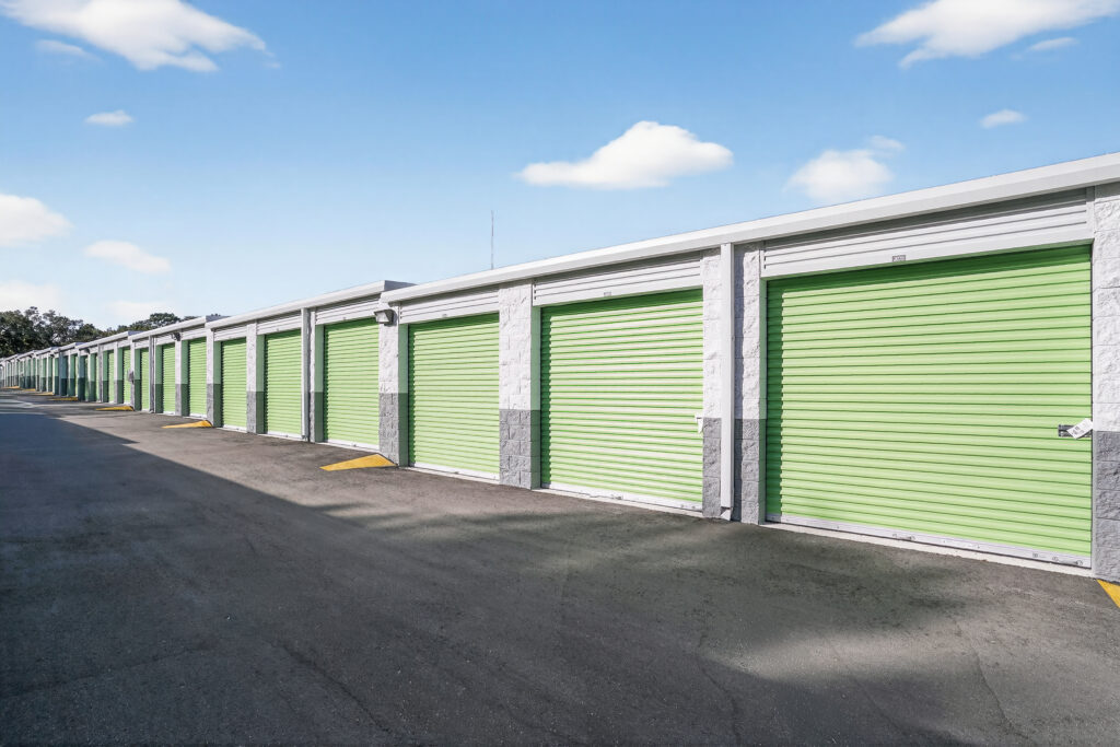 A row of self-storage units with green roll-up doors along an asphalt driveway.