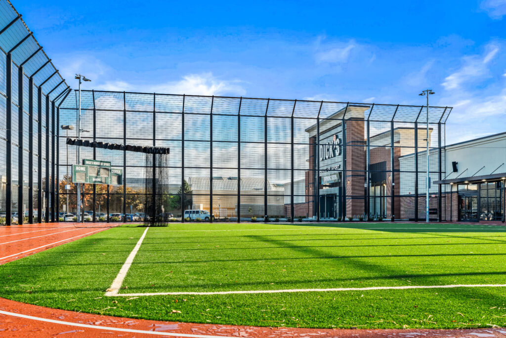 Outdoor athletic field with artificial turf, a scoreboard, and a large net enclosure in front of a Dick’s House of Sport retail store.
