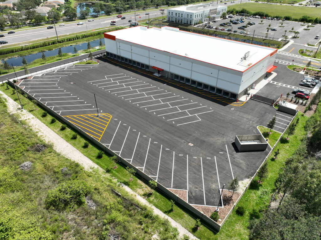 High-angle view of a white, rectangular commercial building with a large empty parking lot in the foreground, situated next to a road and green space.