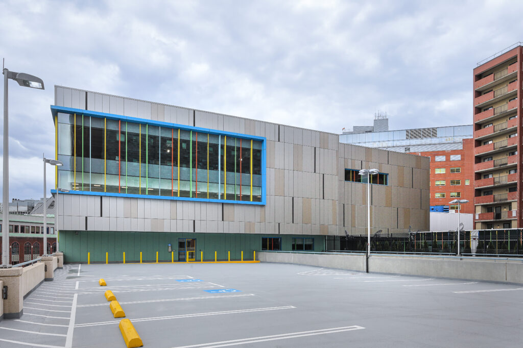 An empty parking lot in front of a modern, multi-story building with a facade of tan panels and a prominent window frame accented with vertical blue, orange, and yellow lines.