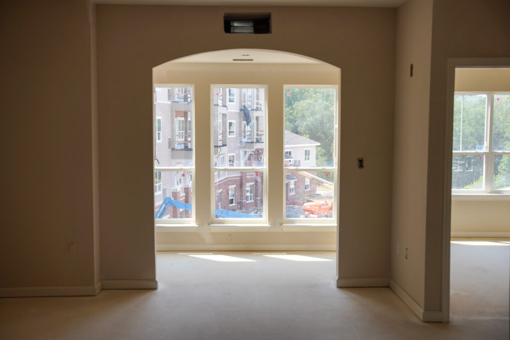 An interior view of a room under construction looking through an arched opening at a multi-story apartment building under construction.