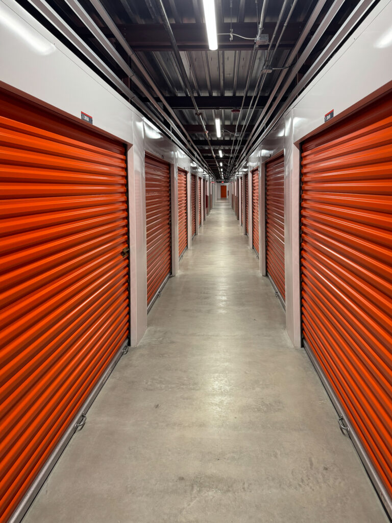 A long, narrow hallway in a self-storage facility lined with rows of closed orange metal rolling doors.