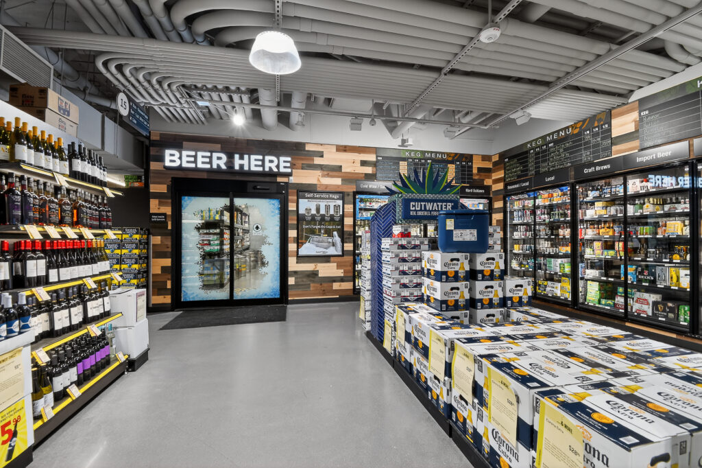 Interior of a beverage store with rows of wine on the left, a walk-in cooler labeled “BEER HERE,” and stacked cases of Corona beer in the foreground.