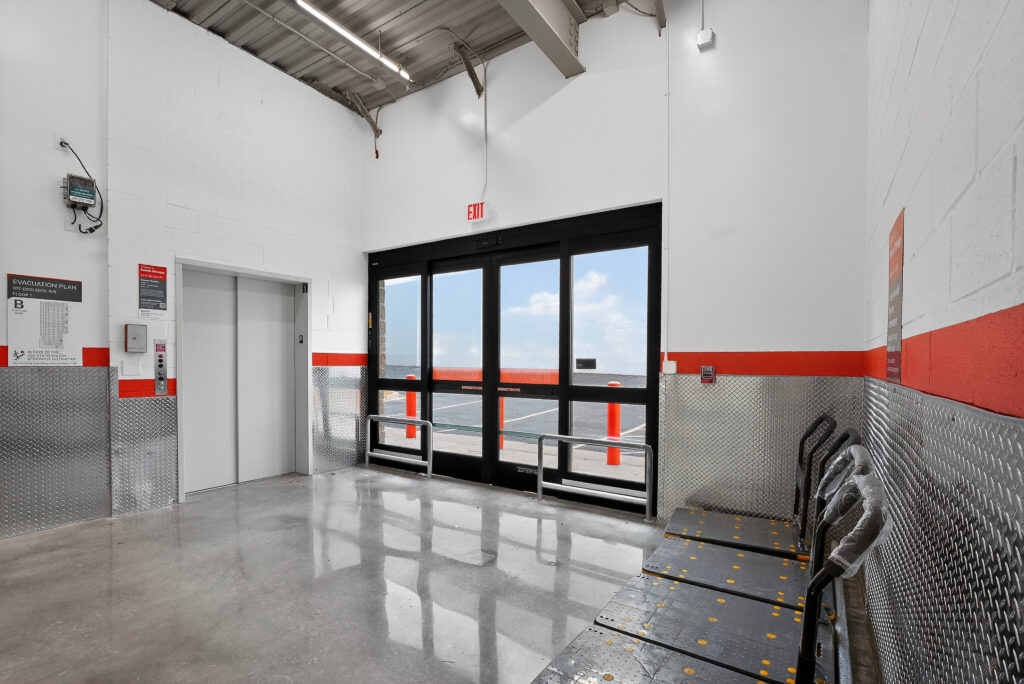 An elevator and a set of sliding glass doors leading to an outdoor parking lot, with a row of plastic-wrapped luggage carts against the wall.