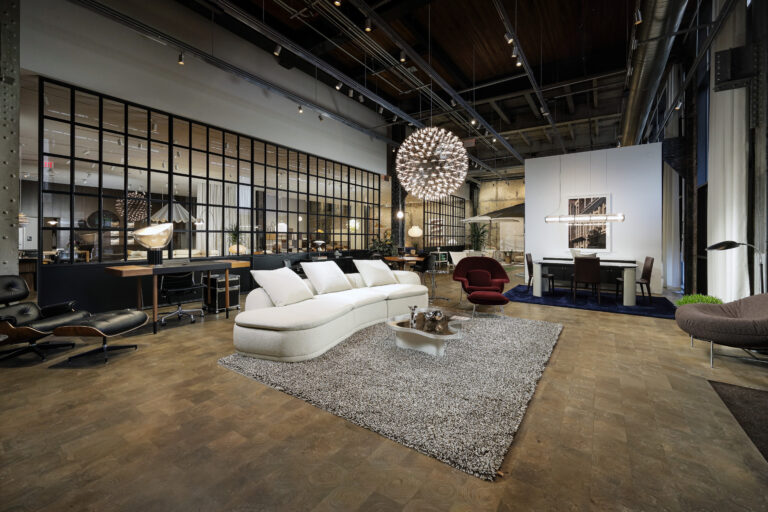 A modern furniture showroom interior featuring a curved white sofa on a shaggy rug, a glass-topped coffee table, a red lounge chair, and a dining set in a high-ceilinged industrial space.