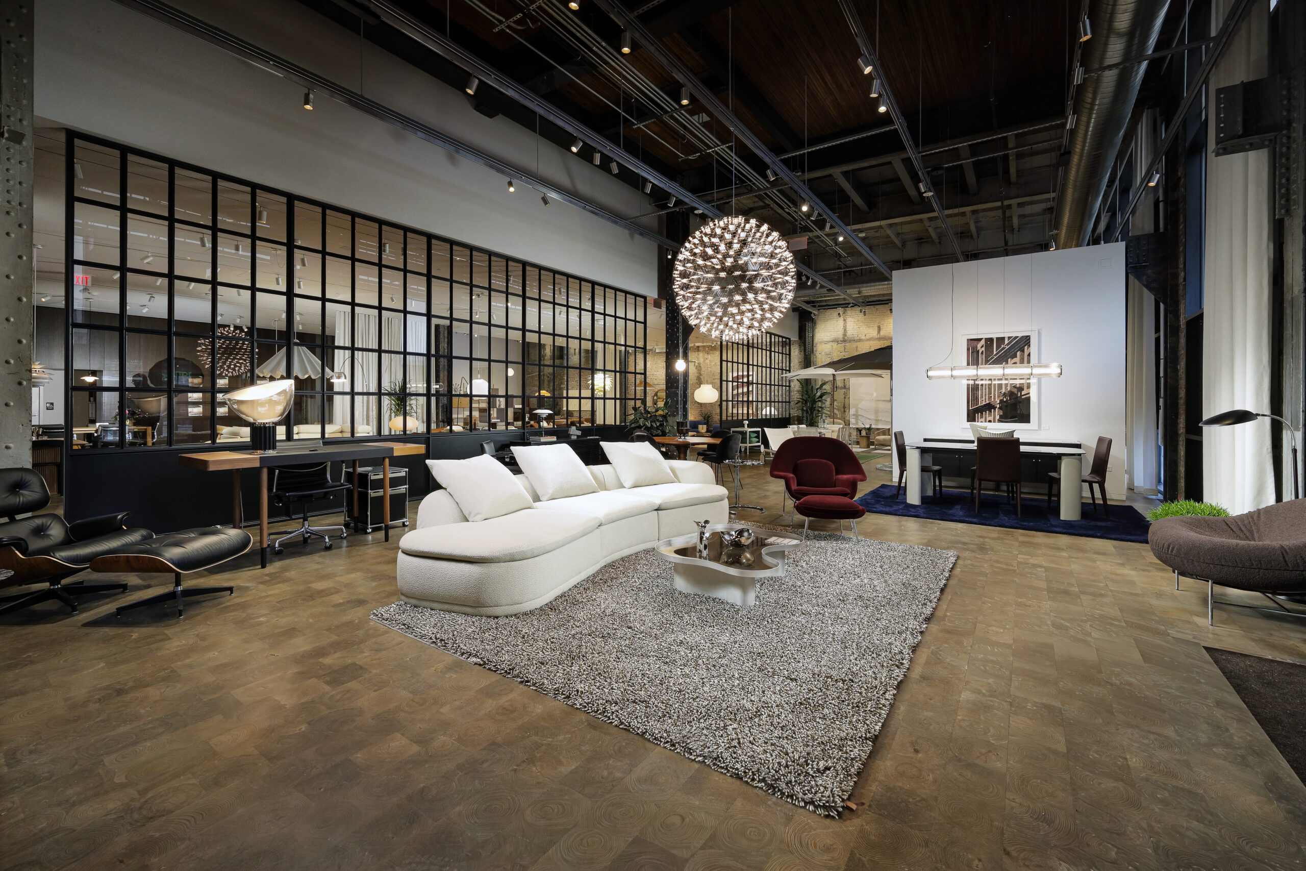 A modern furniture showroom interior featuring a curved white sofa on a shaggy rug, a glass-topped coffee table, a red lounge chair, and a dining set in a high-ceilinged industrial space.