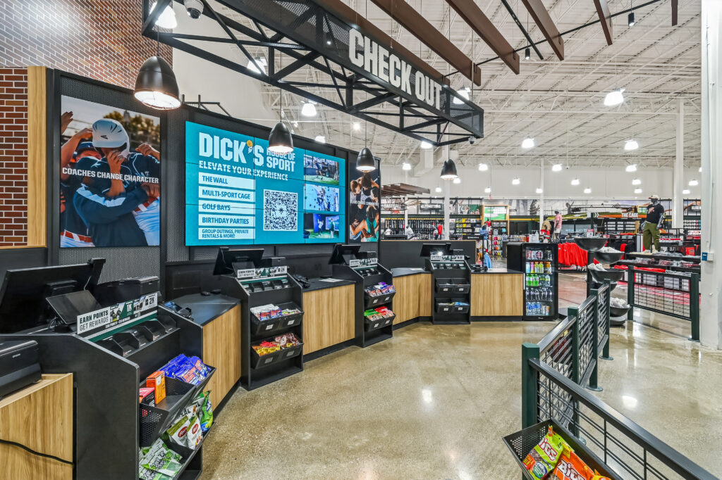 A retail checkout counter at a Dick’s House of Sport store, featuring a digital display promoting store services and a selection of snacks.