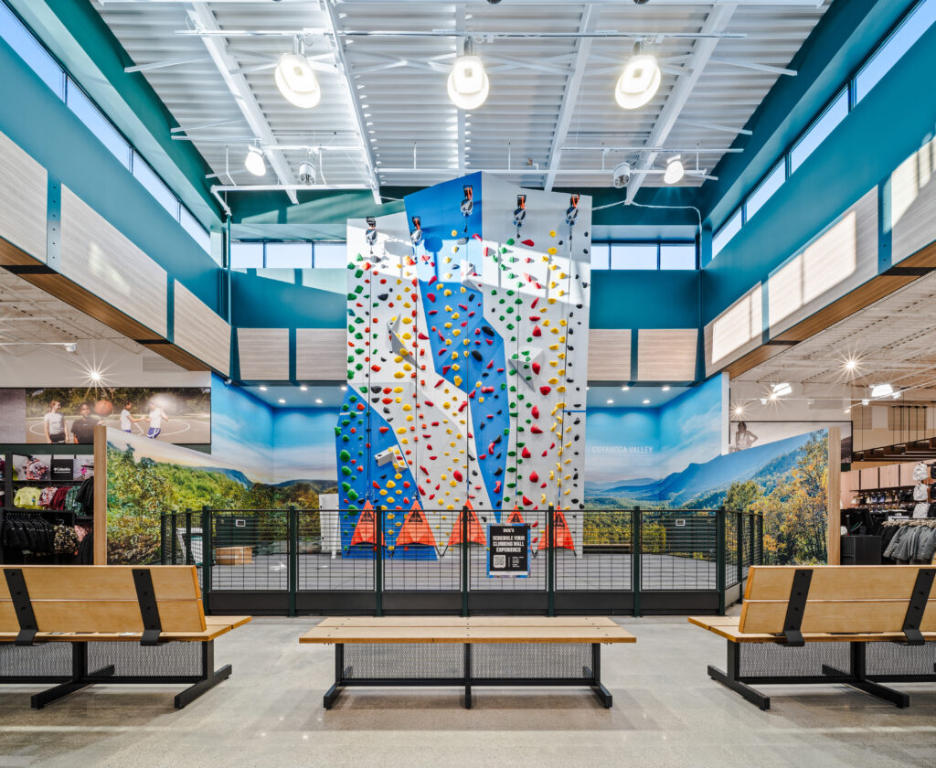 A tall indoor climbing wall with colorful holds stands behind a metal fence, flanked by benches in a retail store.