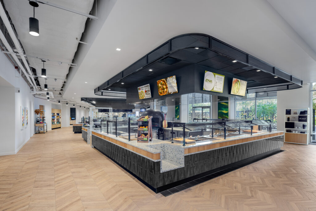 A cafeteria serving station with digital menu boards above a counter, set in a spacious, modern hall with wood-patterned flooring.