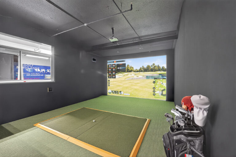 Indoor golf simulator room with a hitting mat, a golf bag in the corner, and a screen displaying a virtual golf course.