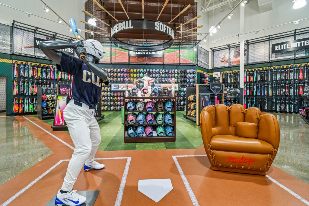 A display in a baseball store featuring a mannequin in a batting stance and a large chair shaped like a baseball glove.