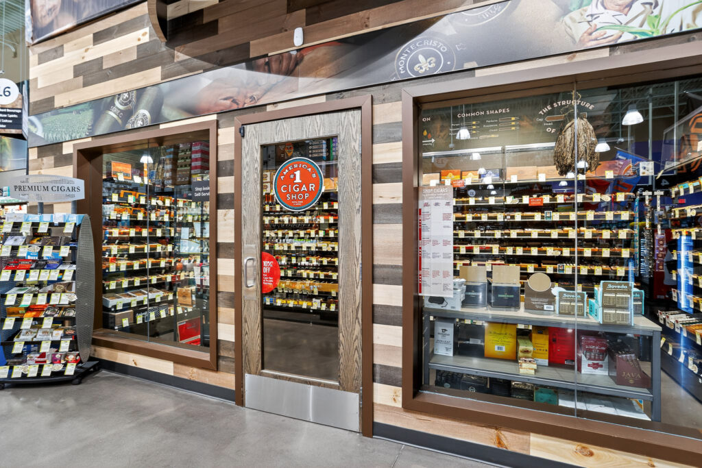 Cigar shop storefront with glass windows and a central door featuring an “America’s #1 Cigar Shop” logo, displaying shelves filled with various cigar boxes.