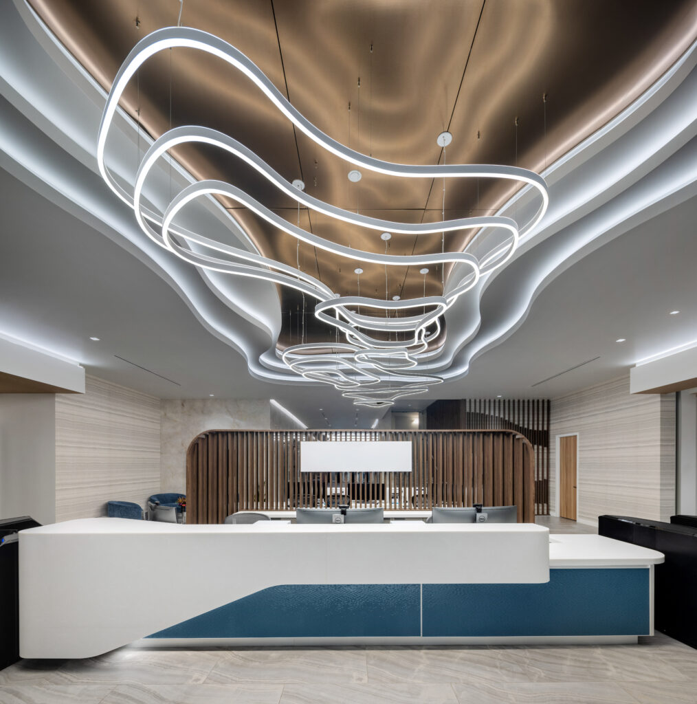 A modern reception area featuring a white and blue curved desk, wooden slat partitions, and a large, abstract white pendant light fixture suspended from a metallic bronze ceiling.